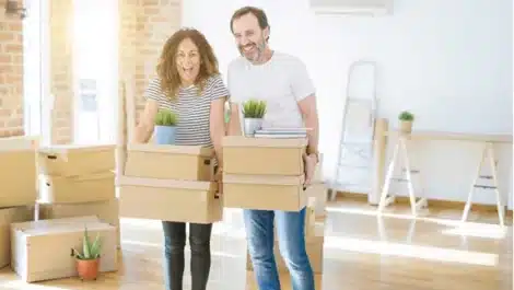 A couple smiling while carrying moving boxes in an empty room.