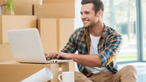 Young man searching on a laptop surrounded by moving boxes.